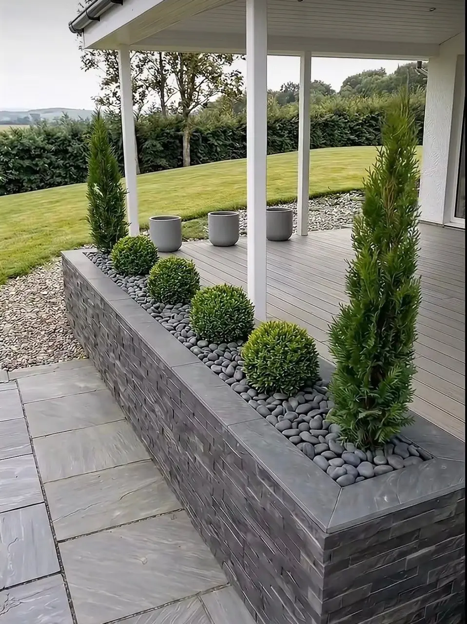 Covered deck with a dark stone planter wall, round shrubs, cypress trees and a stone-paved patio